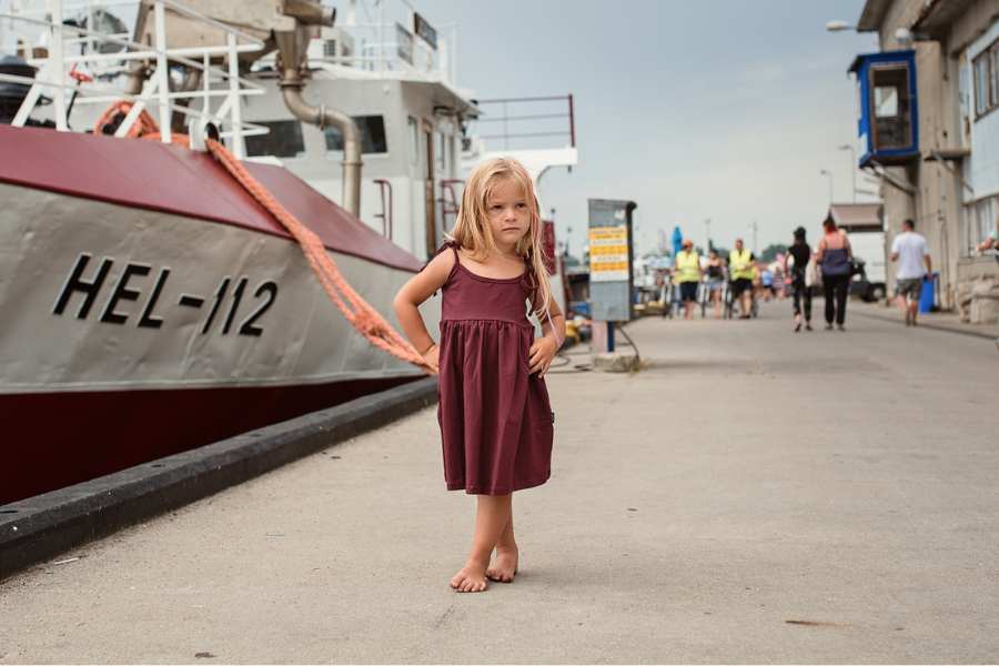 A little girl with long blond hair stands like a model with her hands on her hips next to a ship in a port in the Baltic